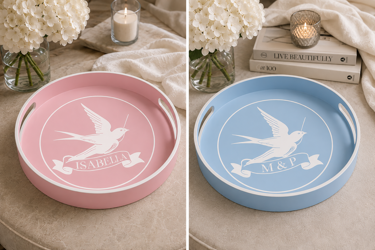Two round serving trays shown side by side in a styled home setting. The left tray is dusty pink with a white bird and banner design reading 'ISABELLA'.