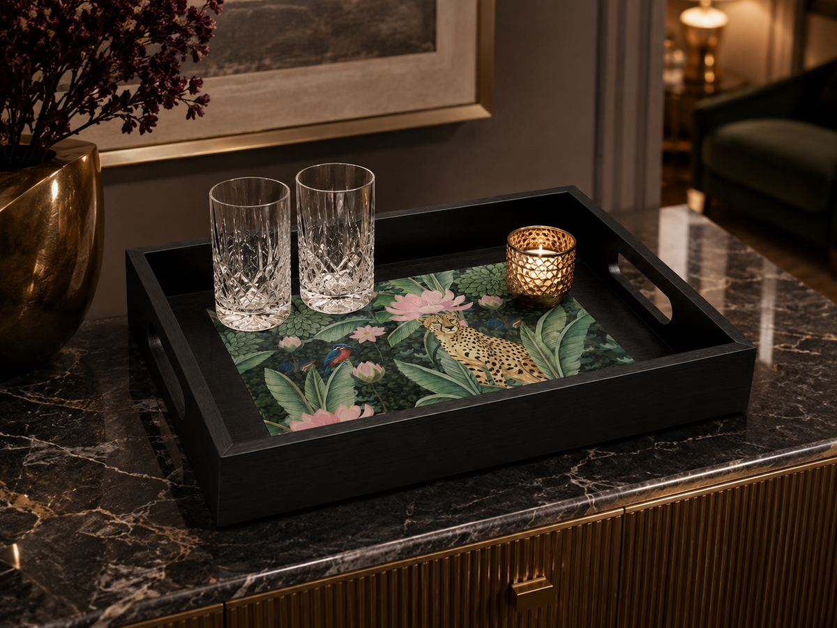 A black rectangular serving tray with handles, featuring a colorful tropical print with green leaves, pink flowers, and a leopard motif, styled on a dark marble countertop with two clear drinking glasses and a gold candle holder.