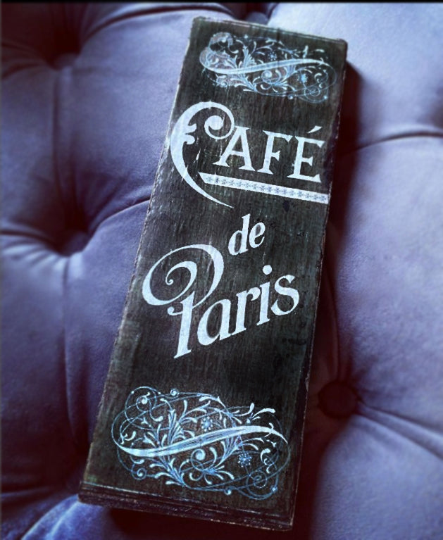 A rectangular dark wooden sign with 'Café de Paris' in white script and ornate flourishes, resting on a tufted purple upholstered surface.