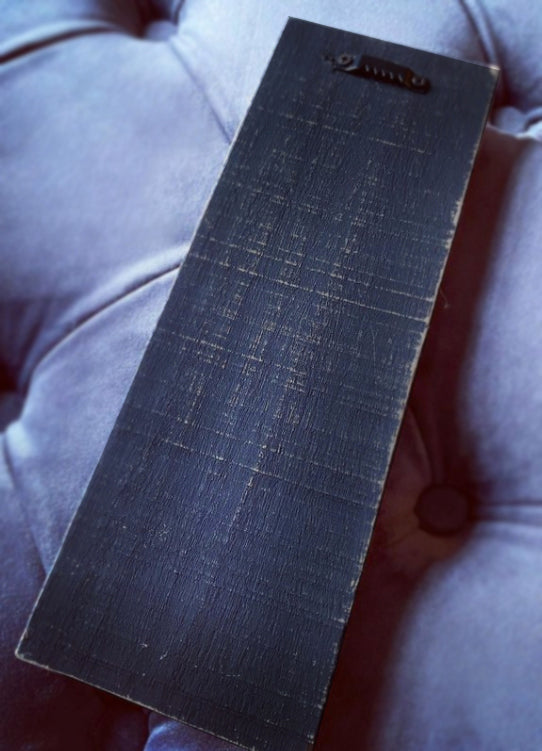 A dark charcoal gray rectangular panel with a brushed metal or textured wood grain finish, featuring a small metal loop at the top, resting on a tufted gray fabric surface.