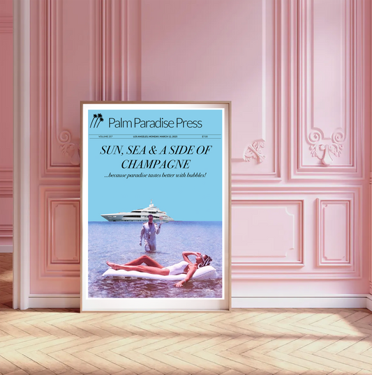 A framed travel poster from Palm Paradise Press leaning against a pink paneled wall on a light wood floor. The poster features a light blue header with the title 'SUN, SEA & A SIDE OF CHAMPAGNE' and a photo of a woman relaxing in the sea on a lilo and a waiter, with a yacht in the background.