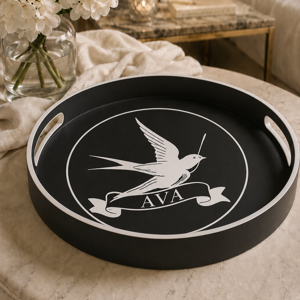 A round black serving tray with white handles and a white printed bird emblem and banner reading 'AVA', styled on a round upholstered ottoman in a warm living room setting with flowers and a lamp in the background.