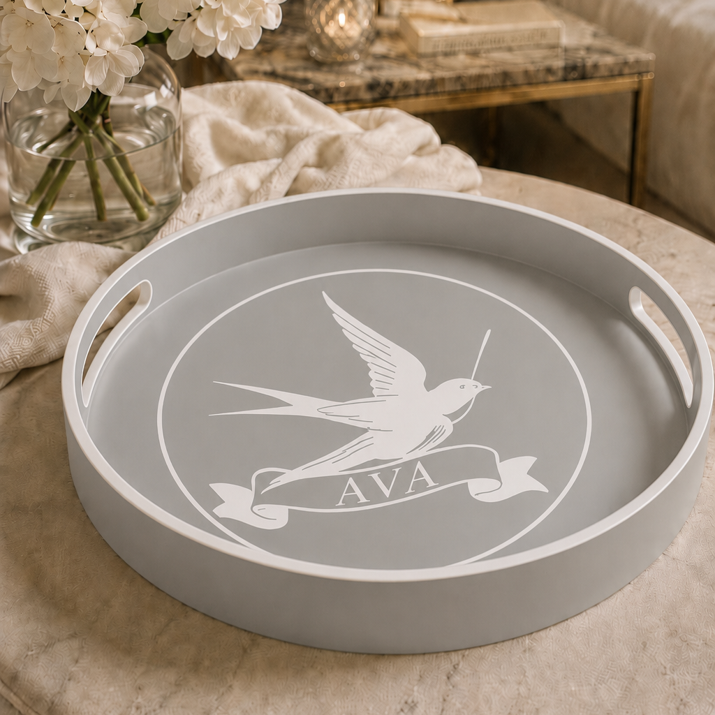 A round grey serving tray with white cutout handles and a white swallow emblem containing the letters 'AVA', displayed on a beige upholstered ottoman in a styled living room setting.
