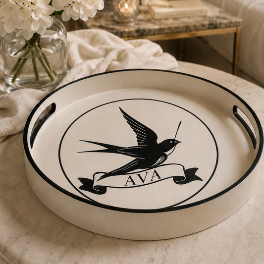 A round white serving tray with black handles and a black printed swallow emblem and banner reading 'AVA', styled on a cream upholstered ottoman in a living room setting.