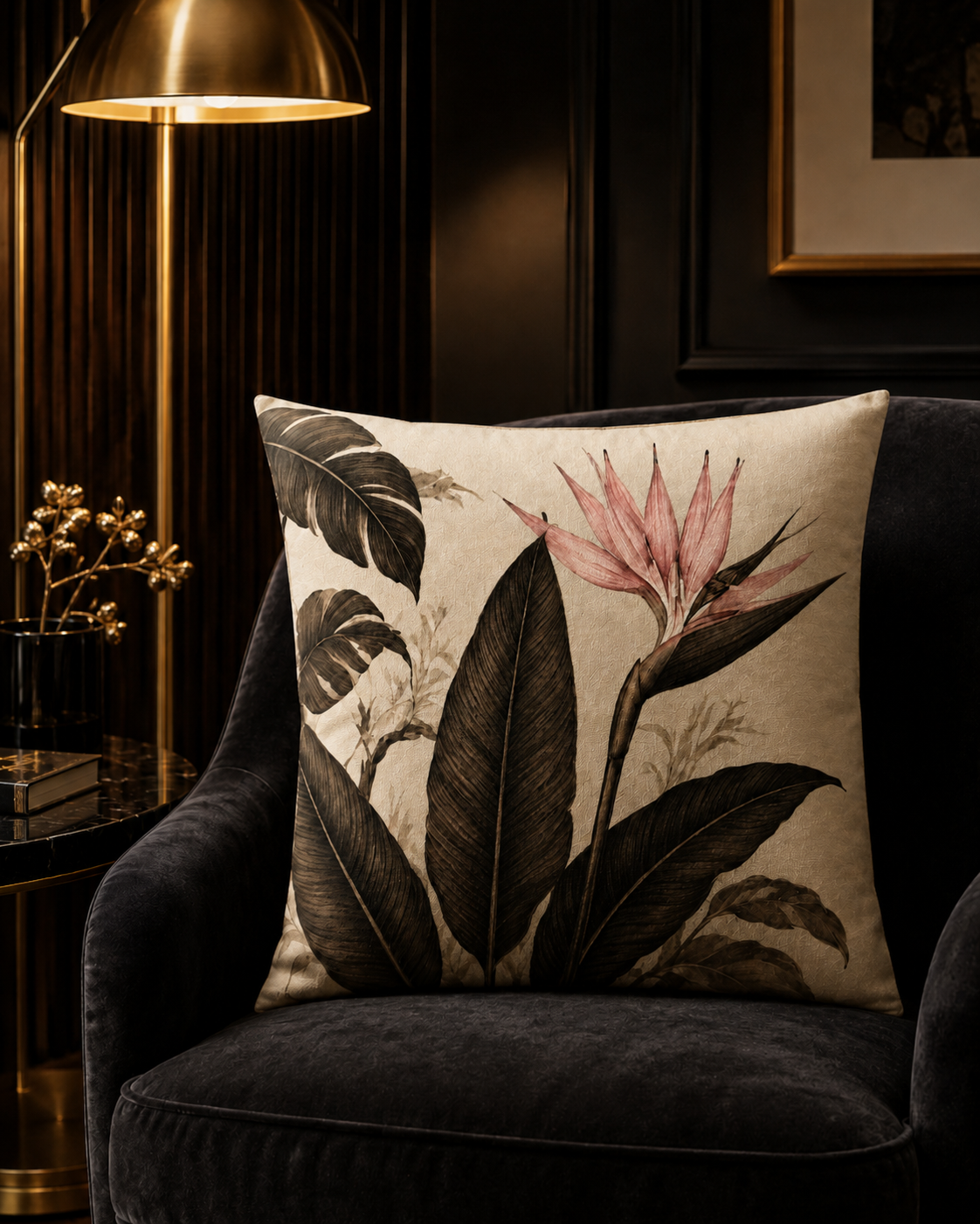 A square decorative throw pillow with a botanical print of dark leaves and a pink bird of paradise flower, resting on a dark velvet armchair in a dimly lit, elegant room.