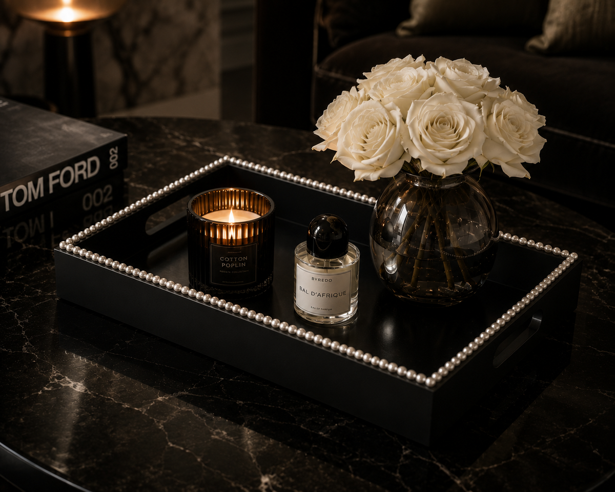 A black rectangular decorative tray with a beaded border sits on a dark marble table, holding a lit candle, a small perfume bottle, and a glass vase filled with white roses. A Tom Ford book is partially visible on the left.