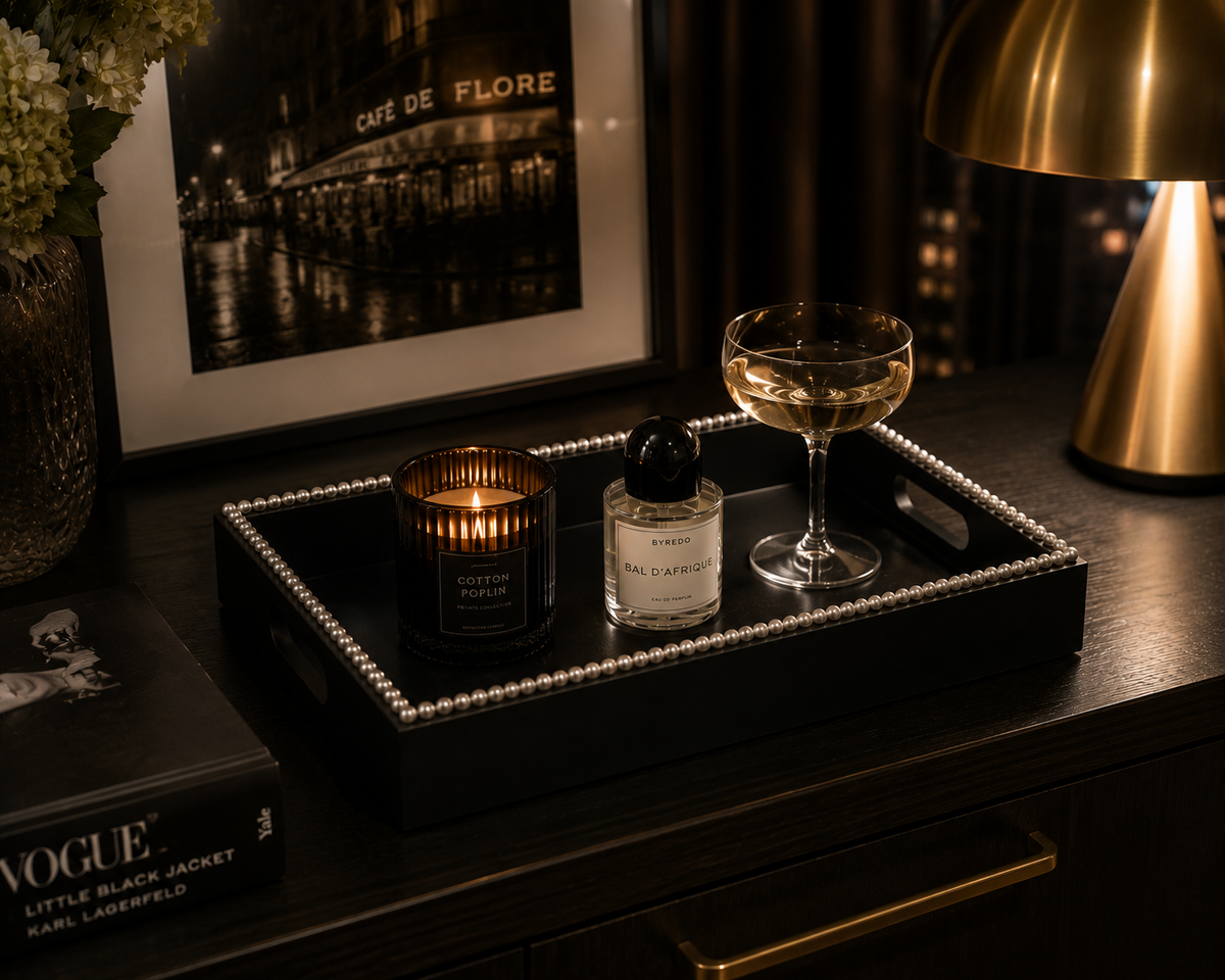 A black decorative tray with a pearl-like border sits on a dark wooden table in a dimly lit room. The tray holds a lit candle, a small perfume bottle, and a coupe glass.