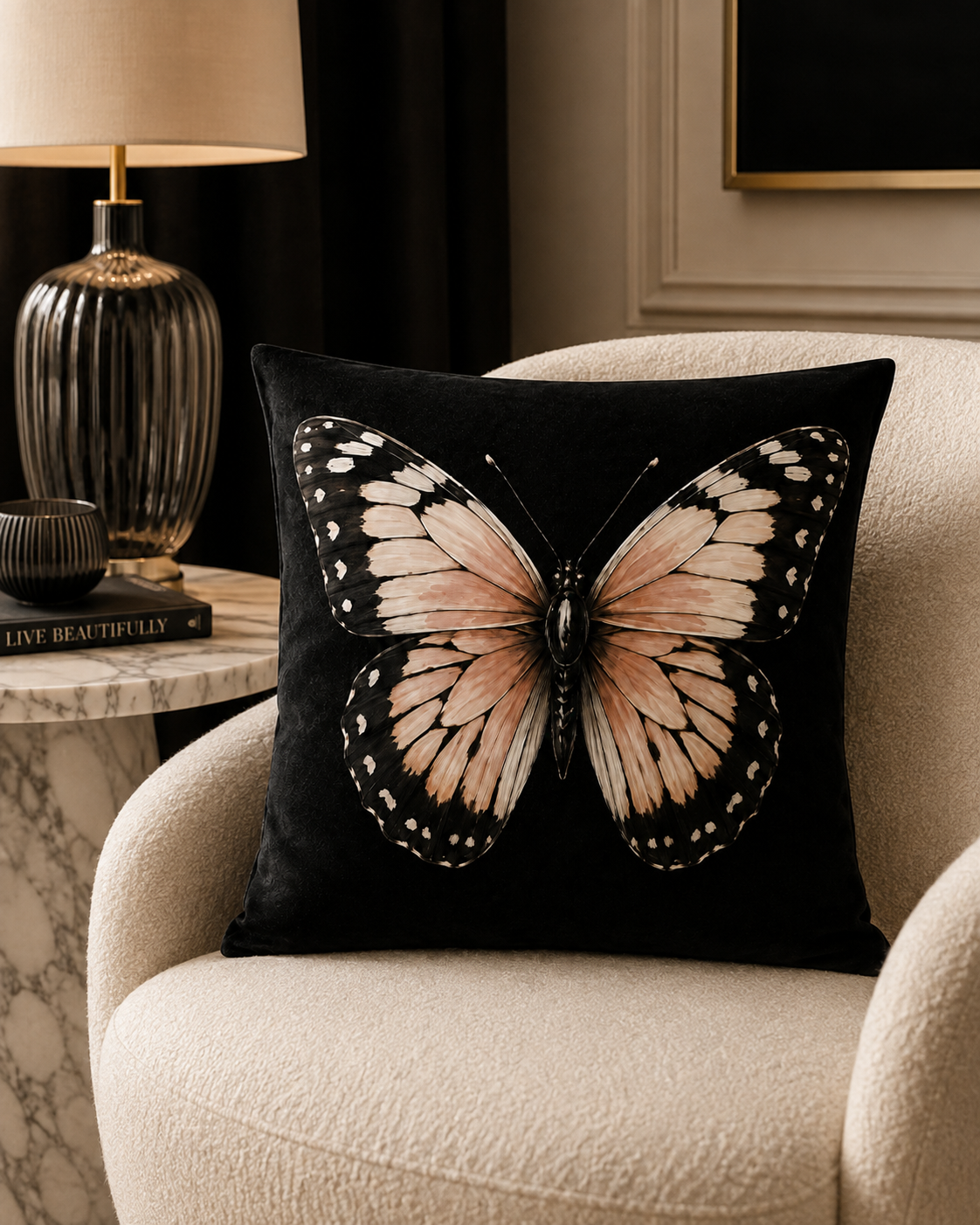 A square black throw pillow featuring a large printed butterfly with pink blush, cream, and black wings, resting on a cream-coloured armchair in a styled living room setting.