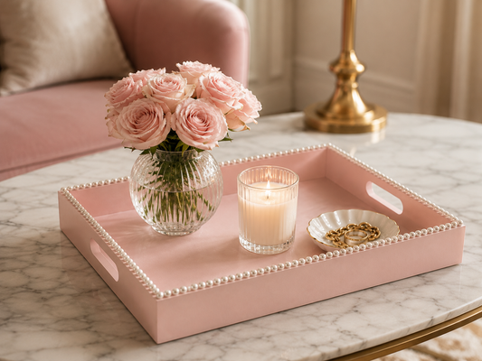 A pink decorative tray with pearl trim sits on a marble table, styled with a vase of pink roses, a lit candle, and a small dish of gold rings in a softly lit interior setting.