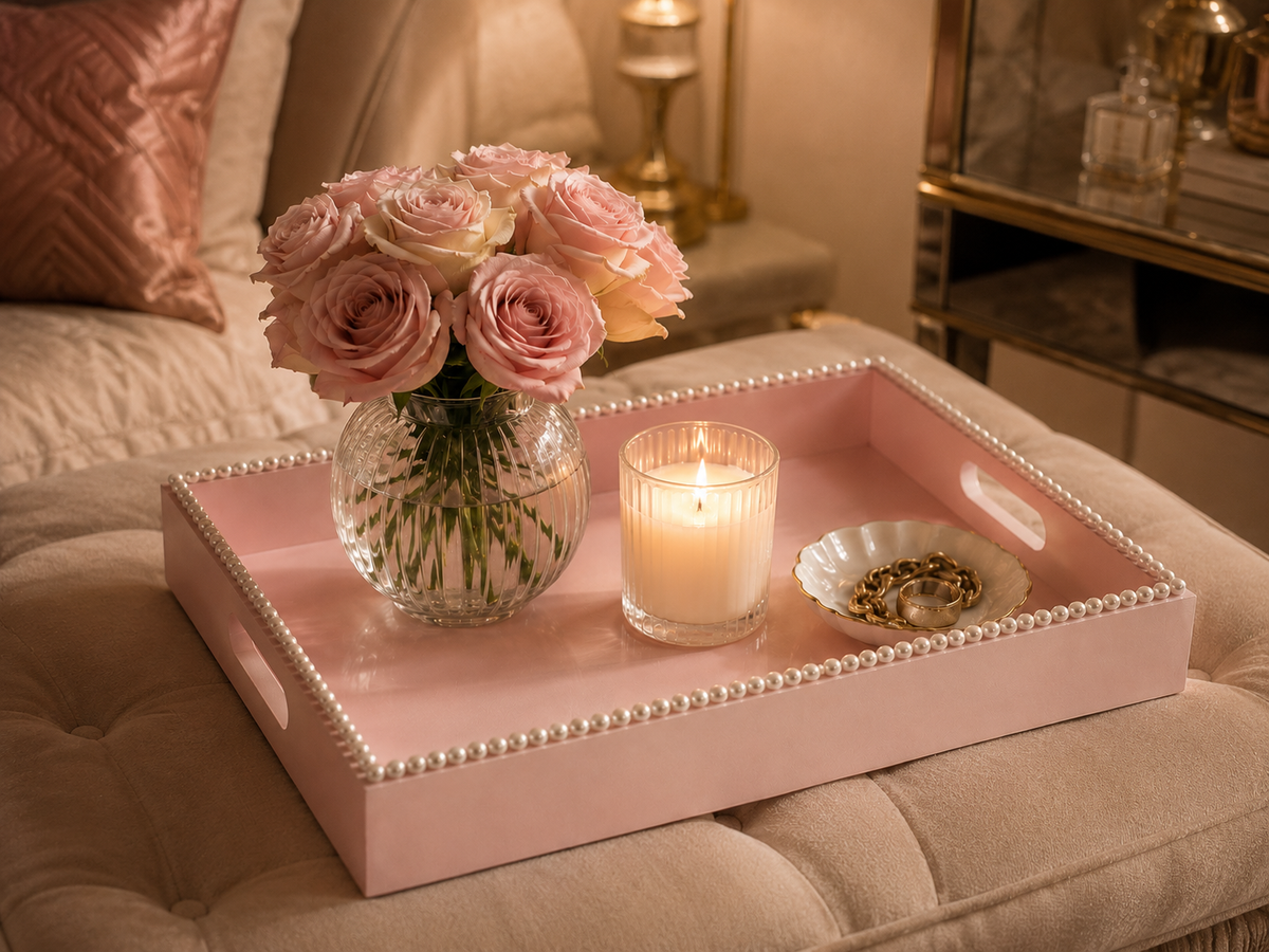 A blush pink pearl edged tray sits on an ottoman with pink roses in a vase, a lit candle and shell tray, in a softly lit luxurious living room.