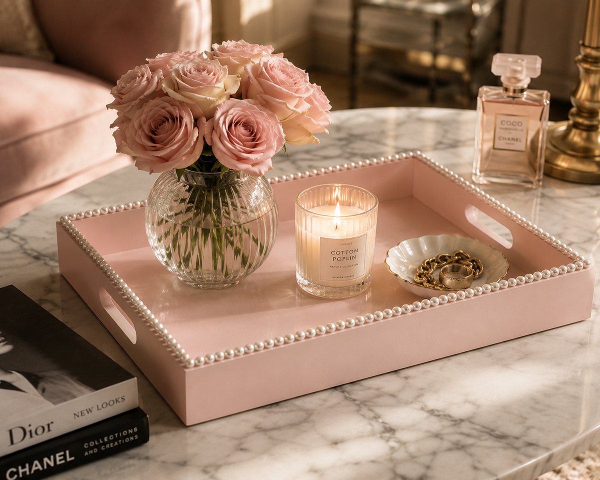 A blush pink rectangular decorative tray with a beaded edge sits on a marble table, styled with a vase of pink roses, a lit candle, a small dish with rings, and perfume bottles in a luxury interior setting.