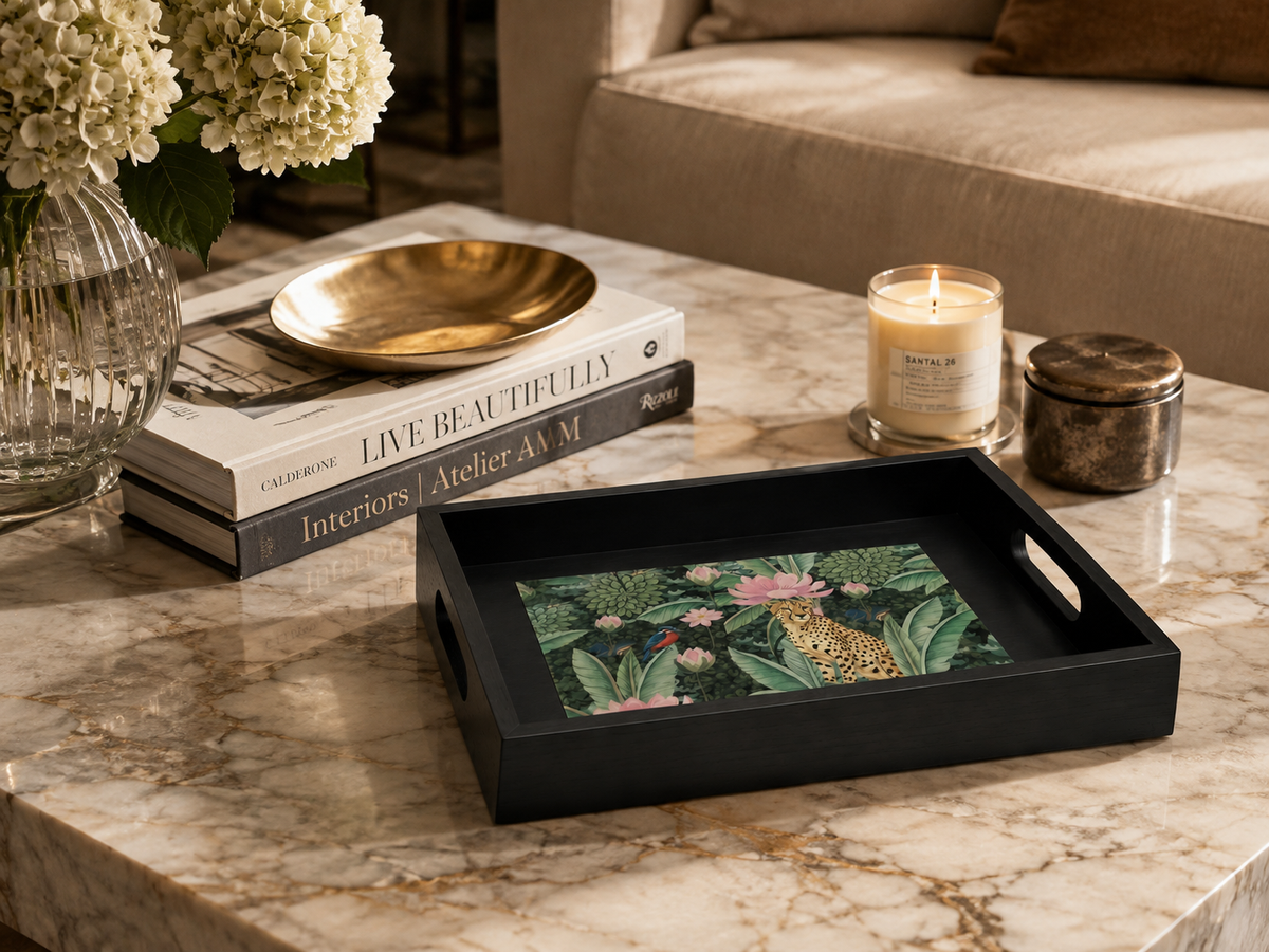 A black rectangular serving tray with cutout handles, featuring a colorful tropical jungle print with a cheetah, is styled on a marble coffee table next to a lit candle, books, and flowers.