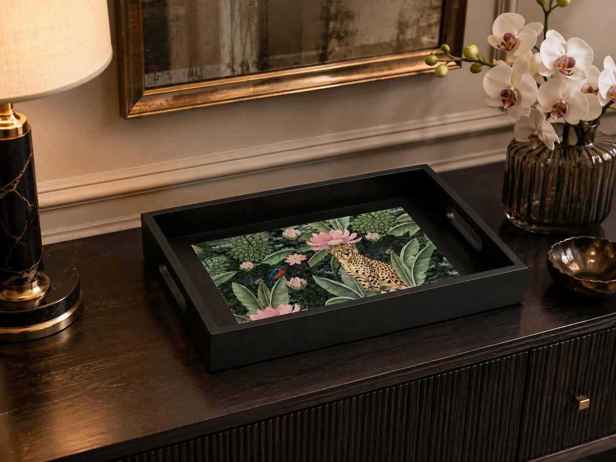 A rectangular black serving tray with cutout handles, featuring a detailed tropical jungle print with a leopard, pink lotus flowers, and green foliage, styled on a dark wood console table next to a lamp and a vase of white orchids.