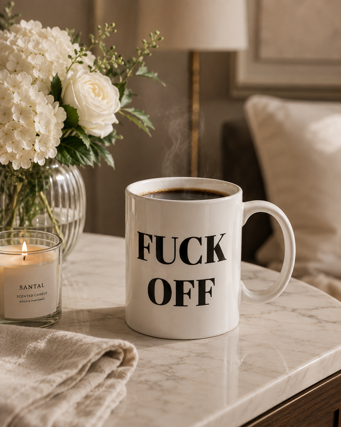 A white ceramic coffee mug with bold black text reading 'FUCK OFF' sits on a marble table in a cozy bedroom setting. The mug has steam rising from the coffee inside, next to a lit candle and white flowers.