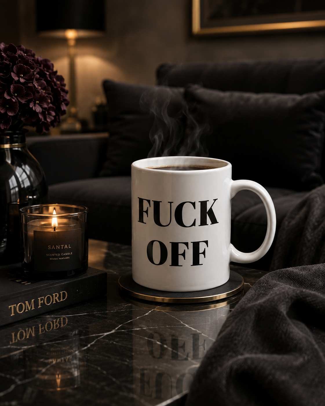 A white ceramic coffee mug with bold black text reading 'FUCK OFF' sits on a gold-rimmed coaster on a dark marble table. The mug is styled in a cozy, dimly lit living room setting with a lit candle, a book, and a dark sofa in the background.