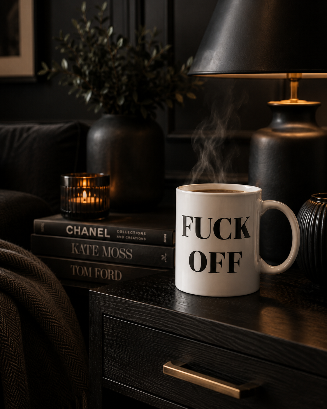 A white ceramic mug with bold black text reading 'FUCK OFF' sits on a dark wooden nightstand in a moody, dimly lit room. Steam rises from the mug, and surrounding props include a lit candle, books, a lamp, and a dark sofa.