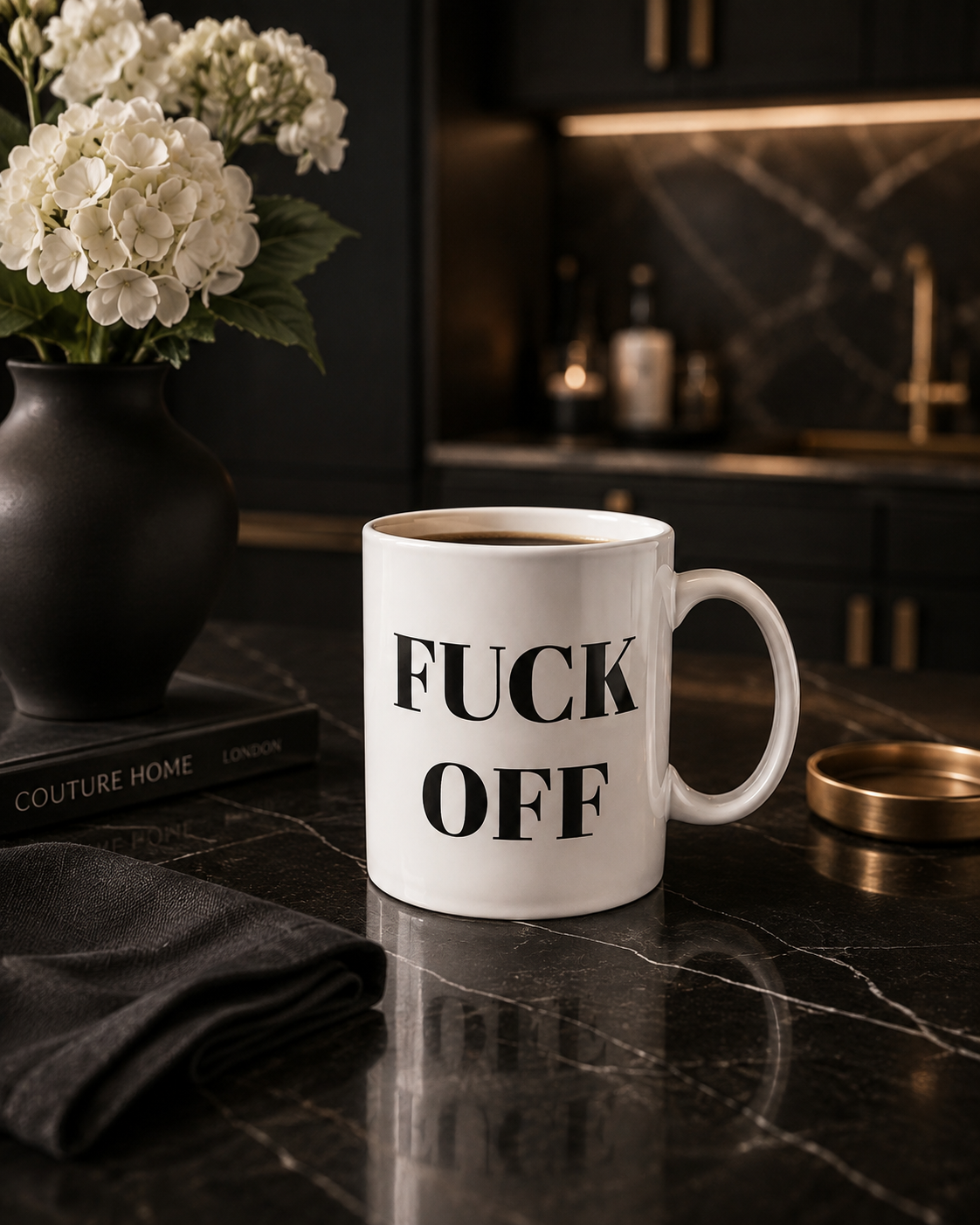 A white ceramic coffee mug with bold black text reading 'FUCK OFF' sits on a dark marble countertop in a modern kitchen setting, next to a black vase of white flowers and a folded dark cloth.