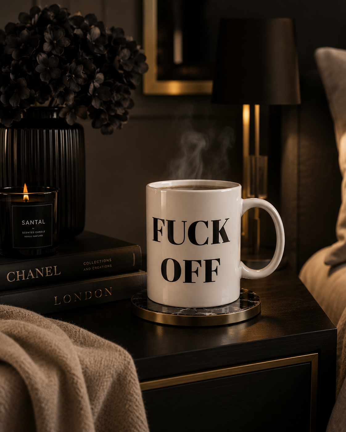A white ceramic mug with bold black text reading 'FUCK OFF' sits on a coaster on a dark bedside table in a moody bedroom setting, with a candle, books, and a lamp in the background.