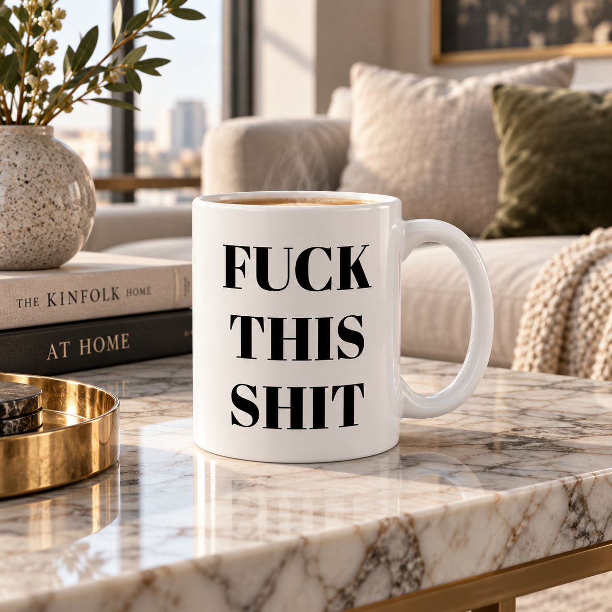A white ceramic mug with bold black text reading 'FUCK THIS SHIT' sits on a marble coffee table in a modern living room setting.