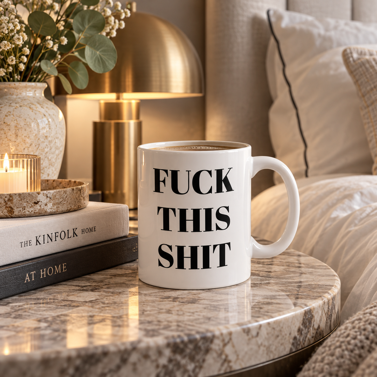 Funny rude coffee mug with black serif typography sits on a marble nightstand in a cozy bedroom setting with a gold lamp, candle, and books in the background