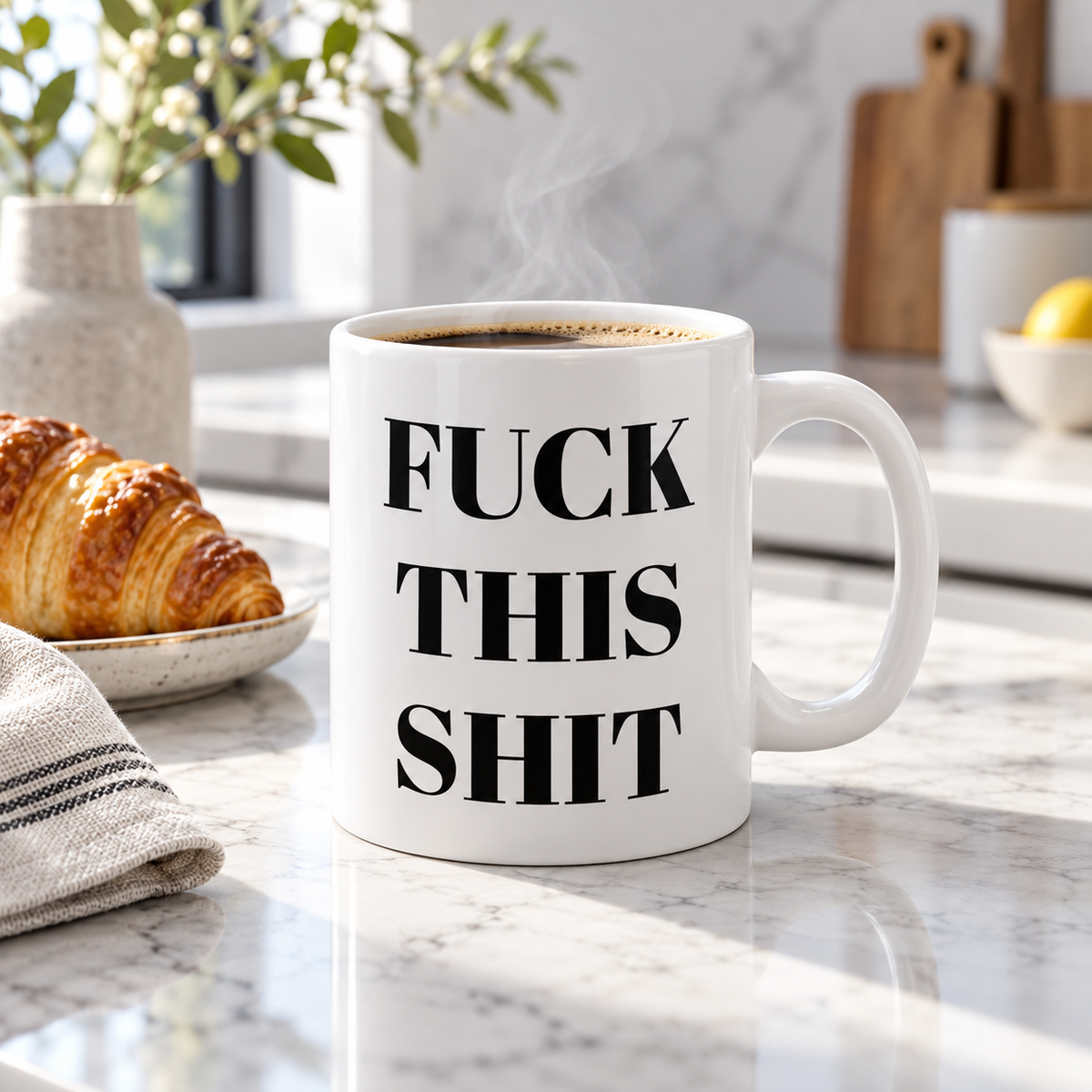 A white ceramic coffee mug with bold black text sits on a marble countertop in a bright kitchen setting, with a croissant and flowers blurred in the background.