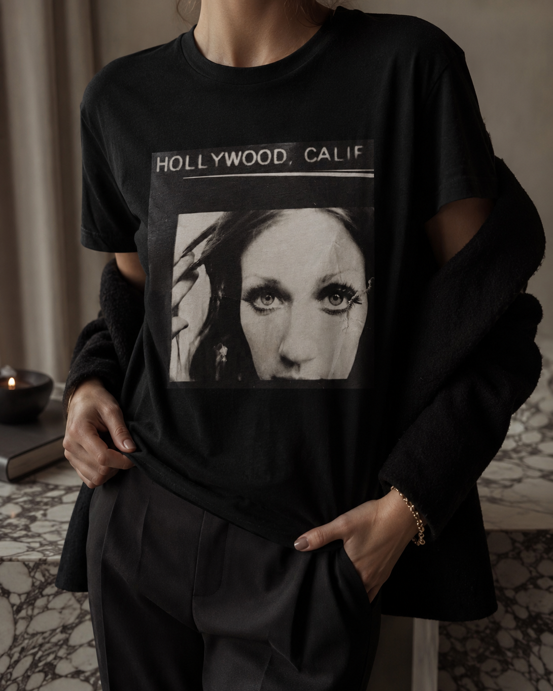A person wearing a black graphic T-shirt with a large black-and-white portrait print and the text 'HOLLYWOOD, CALIF' across the chest, styled with a dark fuzzy cardigan and dark pants in an indoor setting.