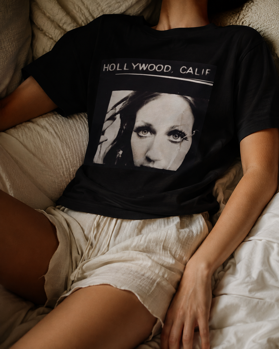 A person reclining on a bed wearing a black graphic T-shirt with the text 'HOLLYWOOD, CALIF' above a black-and-white portrait print, paired with light beige shorts.