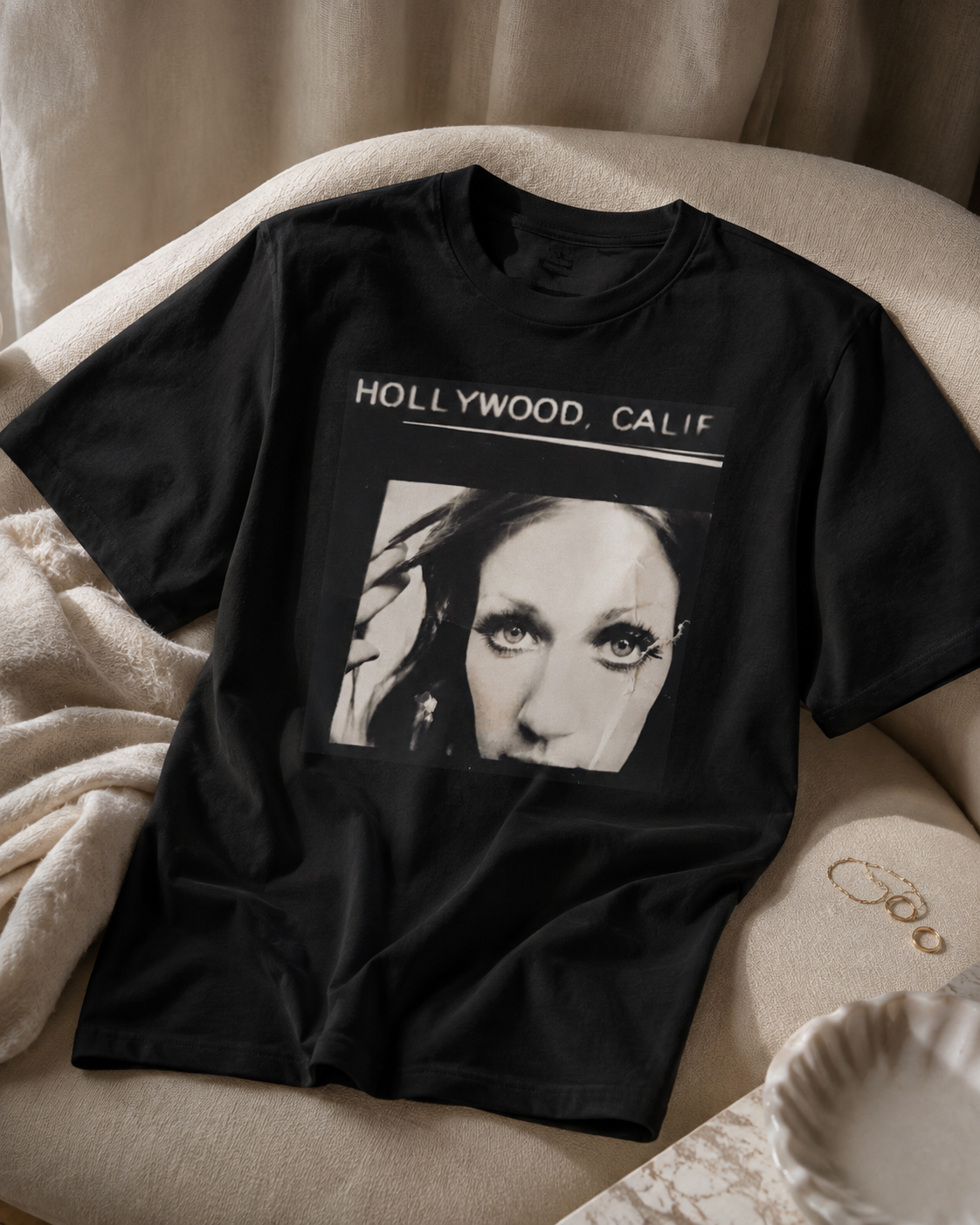 A black short-sleeve graphic T-shirt laid flat on a beige upholstered chair with a soft blanket nearby. The shirt has white text reading 'HOLLYWOOD, CALIF' above a black-and-white portrait print.