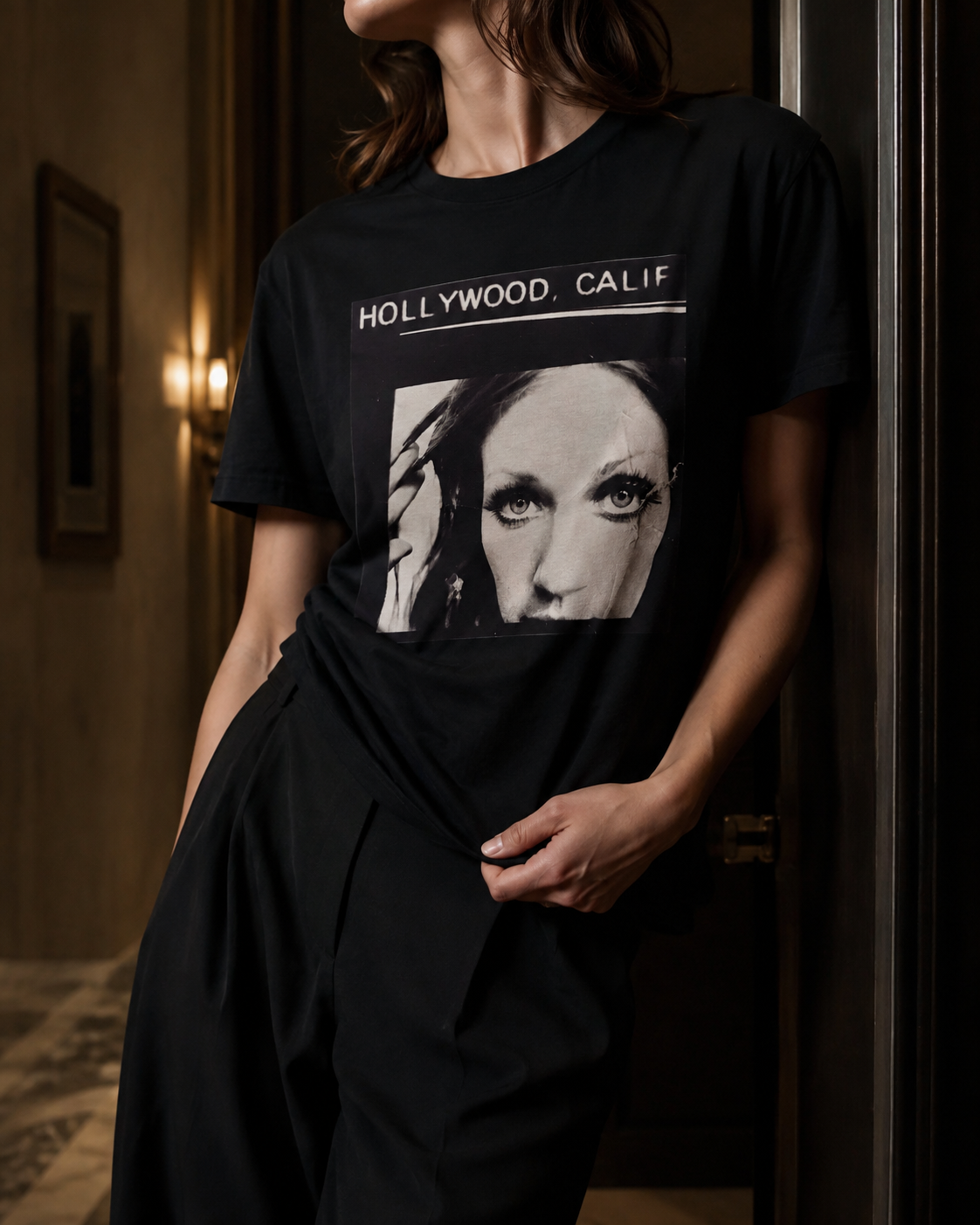 A woman wearing a black graphic T-shirt with a large black-and-white portrait print and the text 'HOLLYWOOD, CALIF' across the chest, posed indoors against a dark doorway.