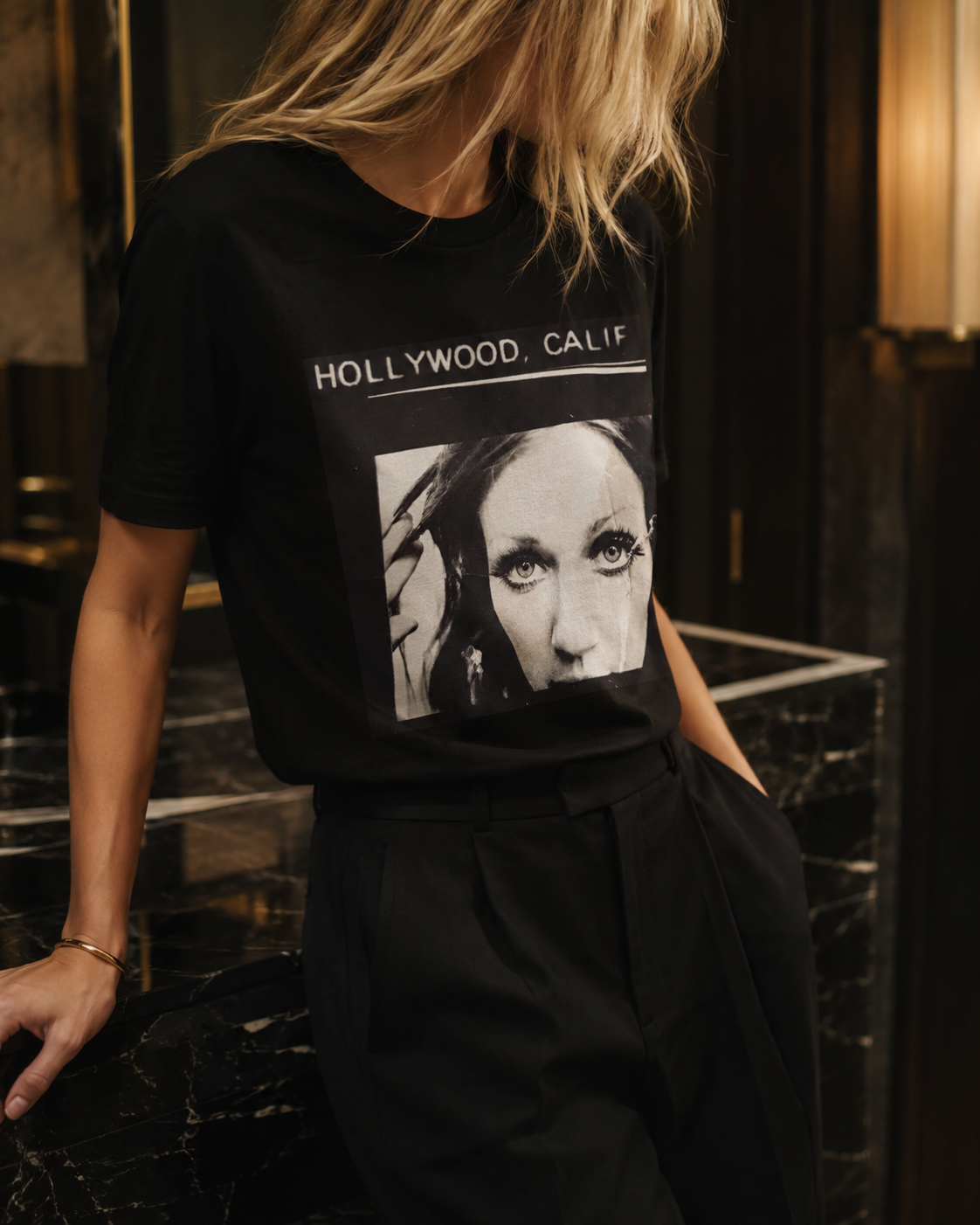 A woman wearing a black graphic t-shirt with a black and white portrait of a woman's face and the text 'HOLLYWOOD, CALIF' printed on the front, paired with black trousers in a dimly lit interior setting.