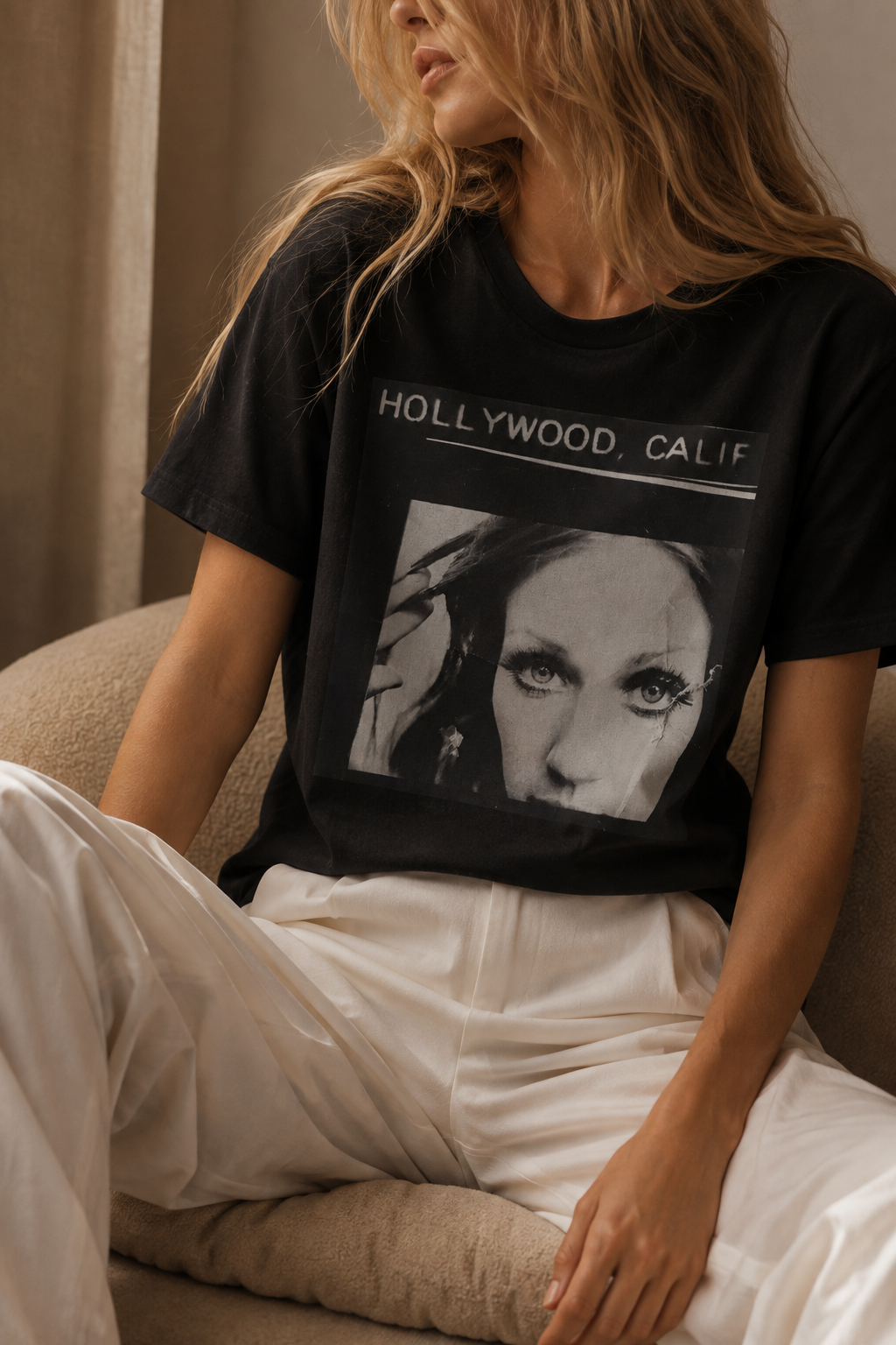 A woman wearing a black graphic t-shirt with a black and white portrait of a woman's face and the text 'HOLLYWOOD, CALIF' across the chest, paired with white pants, sitting on a beige couch.