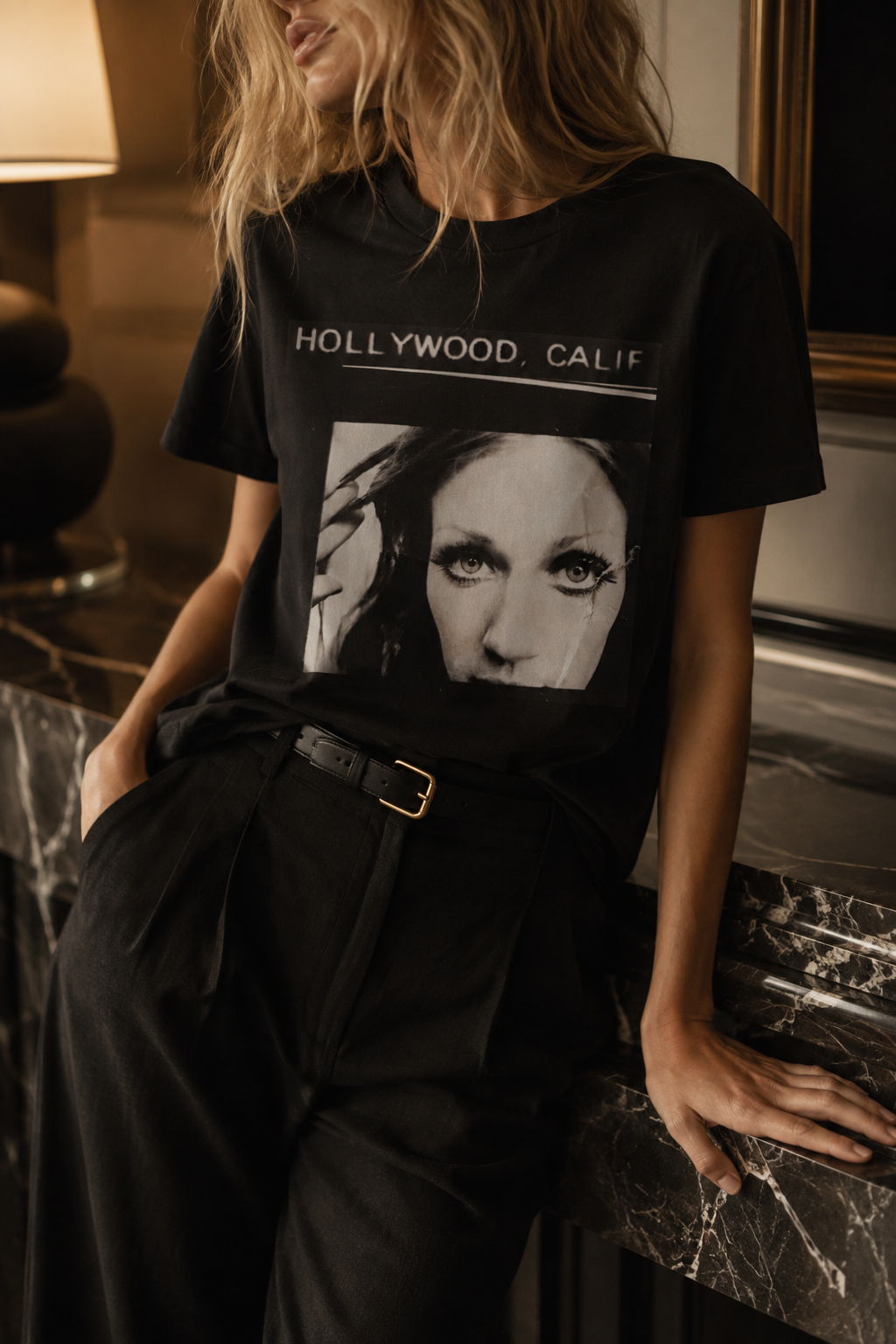 A woman wearing a black graphic t-shirt with 'HOLLYWOOD, CALIF' and a black-and-white portrait print, paired with black pants in a warm-toned indoor setting.