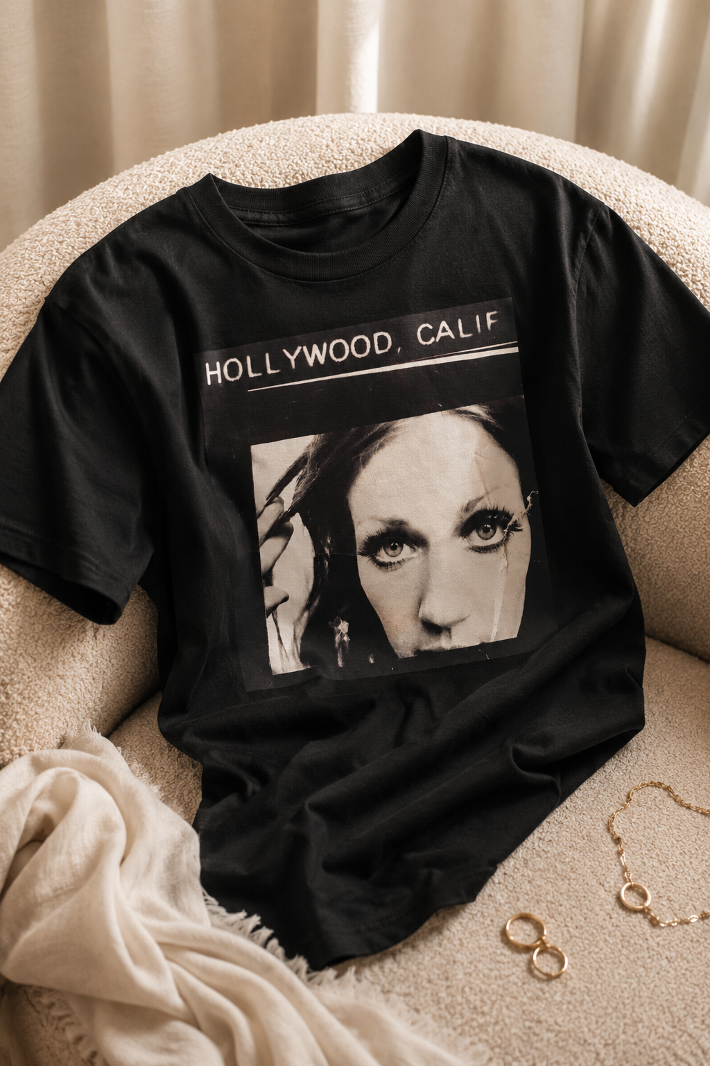 A black graphic t-shirt featuring a black and white portrait of a woman with the text 'HOLLYWOOD, CALIF' printed above, styled on a beige textured armchair with a cream throw blanket and gold rings nearby.
