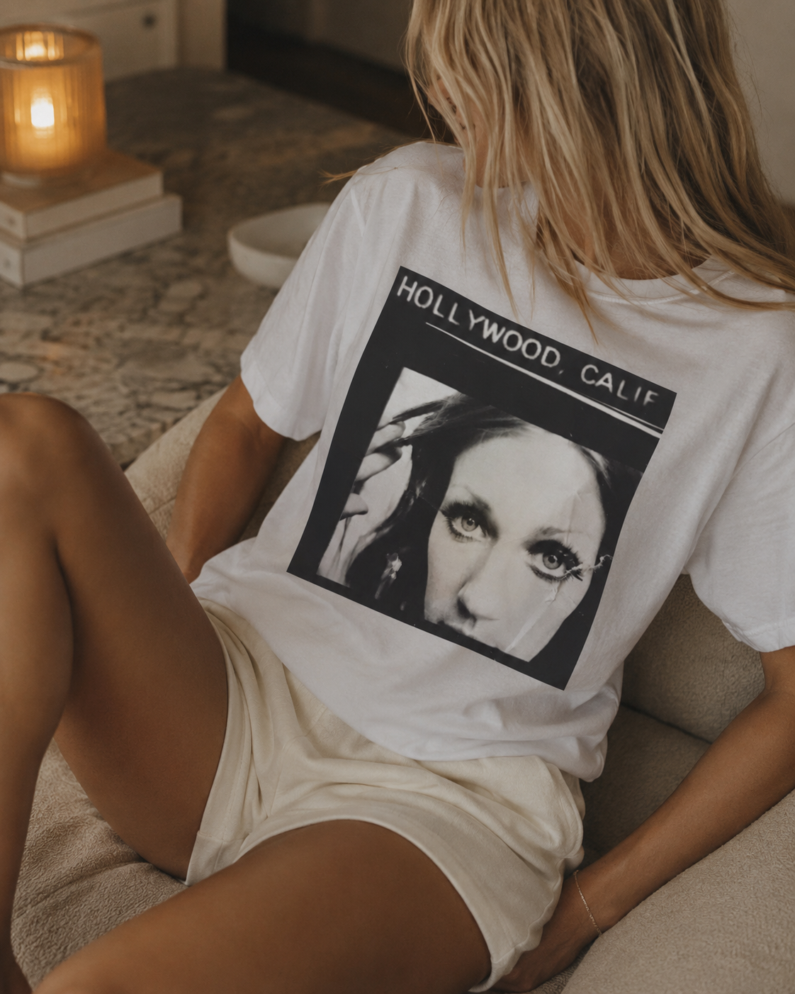 A woman wearing a white graphic t-shirt with a black and white 'Hollywood, Calif.' portrait print, sitting on a beige couch in a cozy room setting.