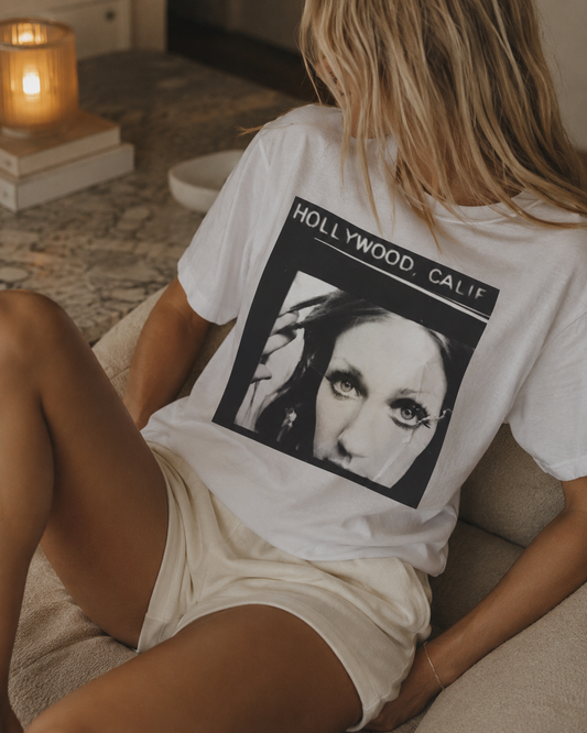 A woman wearing a white graphic t-shirt with a black and white 'Hollywood, Calif.' portrait print, sitting on a beige couch in a cozy room setting.