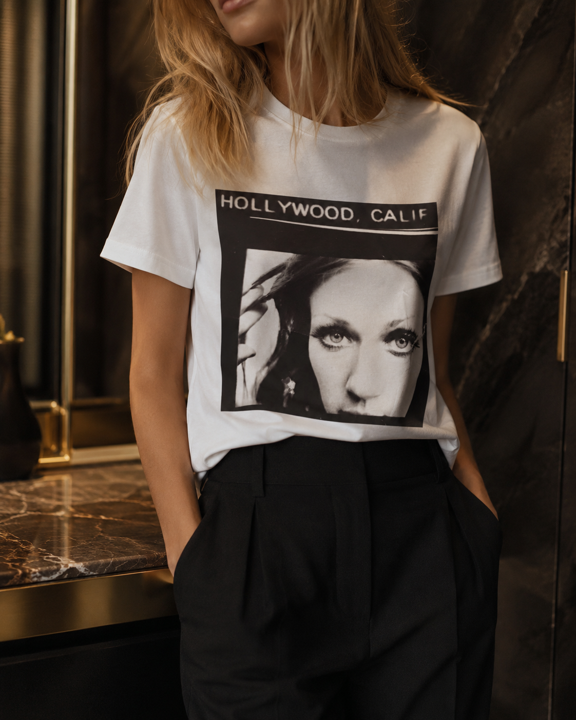A woman wearing a white graphic t-shirt with a black and white print of a face and the text 'HOLLYWOOD, CALIF', paired with black trousers, standing in a dark interior setting.