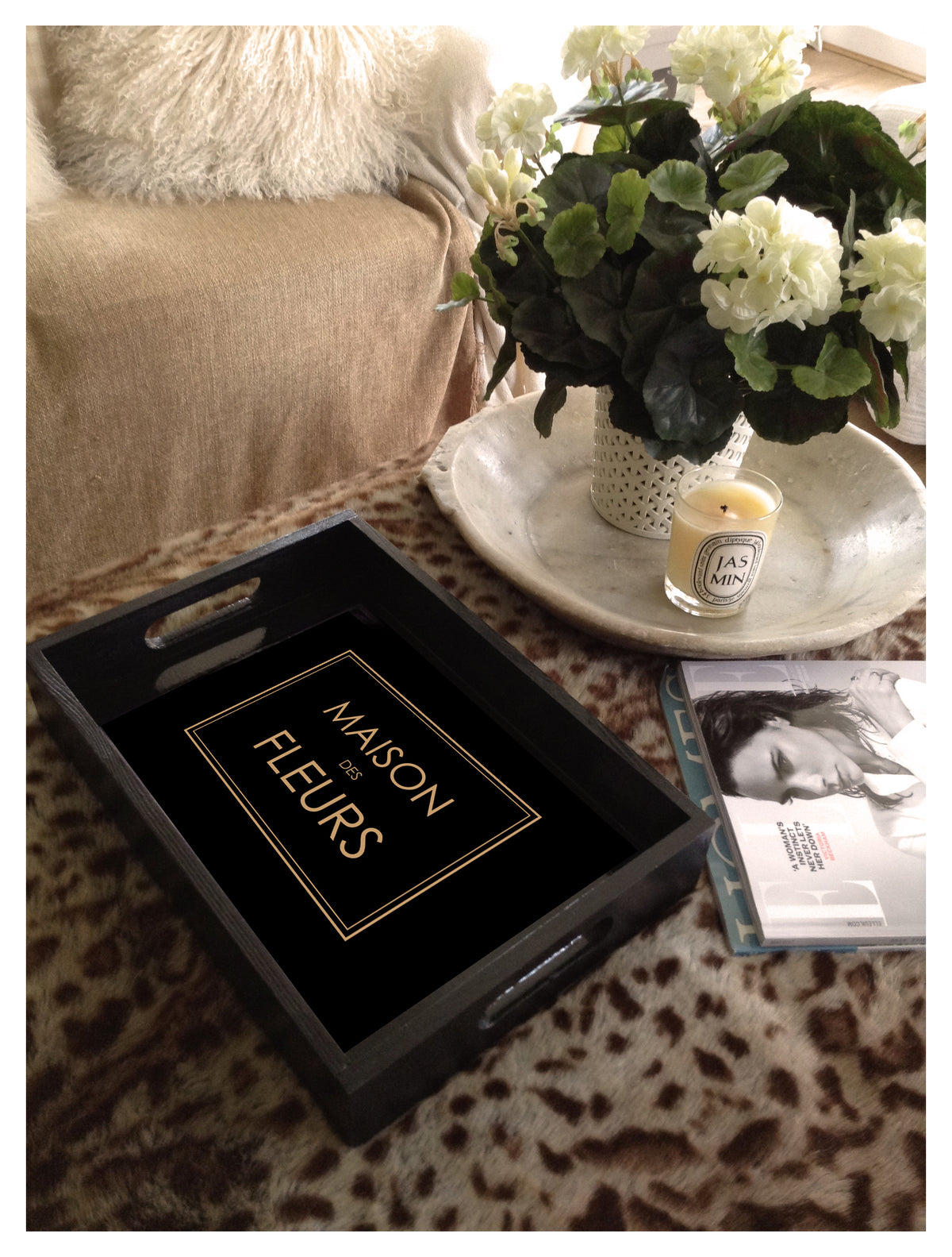 A black rectangular decorative tray with cutout handles and a gold-framed sign reading 'MAISON DES FLEURS' placed on a leopard-print surface. The scene includes a beige sofa, a white tray with flowers, a candle, and a magazine in a styled indoor setting.