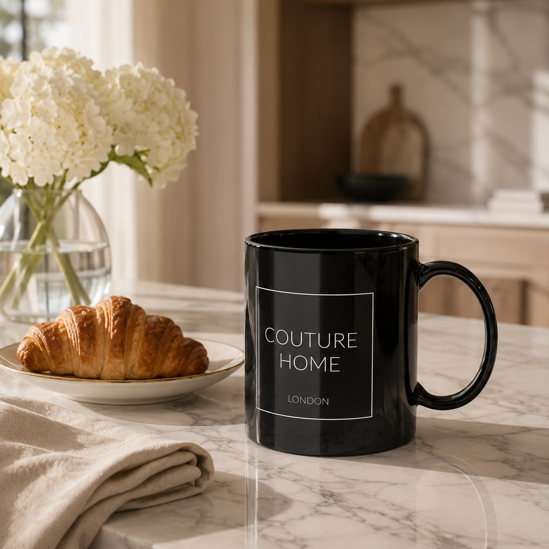 A black ceramic mug with white text reading 'COUTURE HOME LONDON' sits on a marble kitchen counter next to a croissant on a plate and a vase of white flowers.