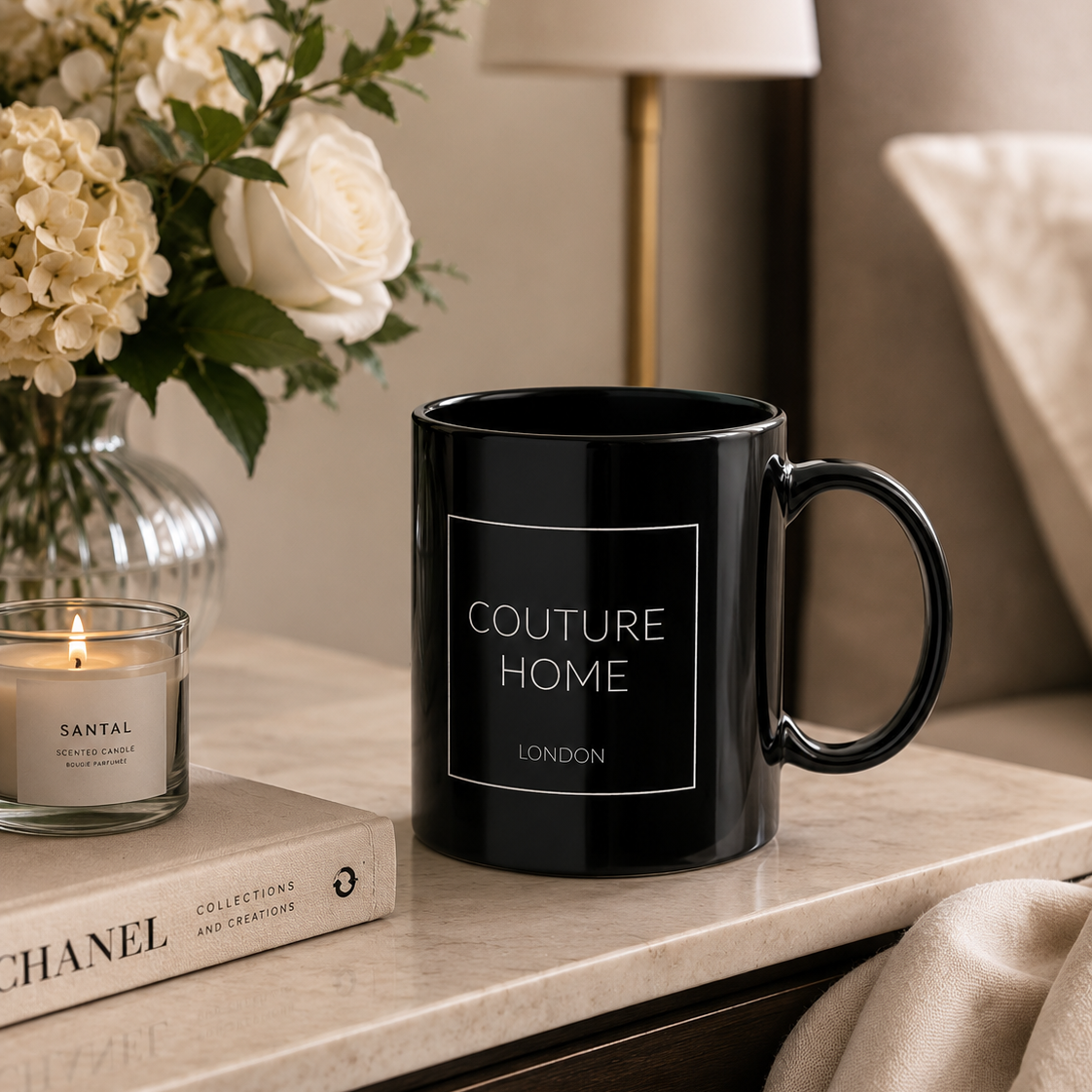 A glossy black ceramic mug with the words 'COUTURE HOME LONDON' printed in white on the front, styled on a marble bedside table next to a candle, a book, and a vase of flowers.
