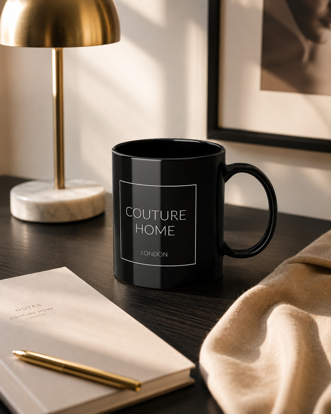 A glossy black ceramic mug with a white square border and the text 'COUTURE HOME LONDON' printed on the side, set on a dark wooden desk next to a gold lamp, notebook, and beige fabric.