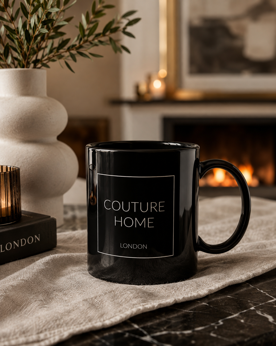 A glossy black ceramic mug with a white printed label reading 'COUTURE HOME LONDON' sits on a beige linen runner in a cozy indoor setting near a fireplace. A white vase with greenery and a candle are visible in the background.