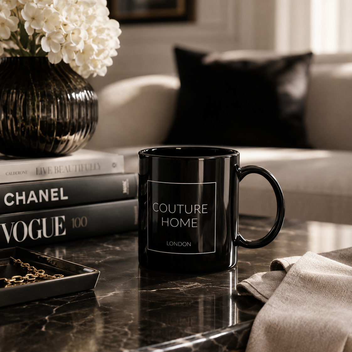 A glossy black ceramic mug with the words 'COUTURE HOME LONDON' printed in white on the front, placed on a dark marble coffee table in a styled living room setting.