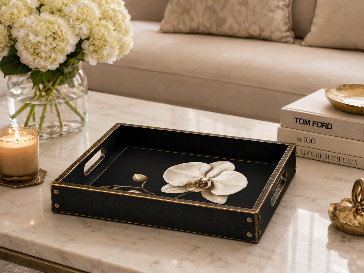 A rectangular black serving tray with gold trim and cutout handles sits on a white marble coffee table in a styled living room. The tray features a black-and-white orchid print in the center and is surrounded by decorative items including a candle, a vase of white flowers, books, and a small gold object.