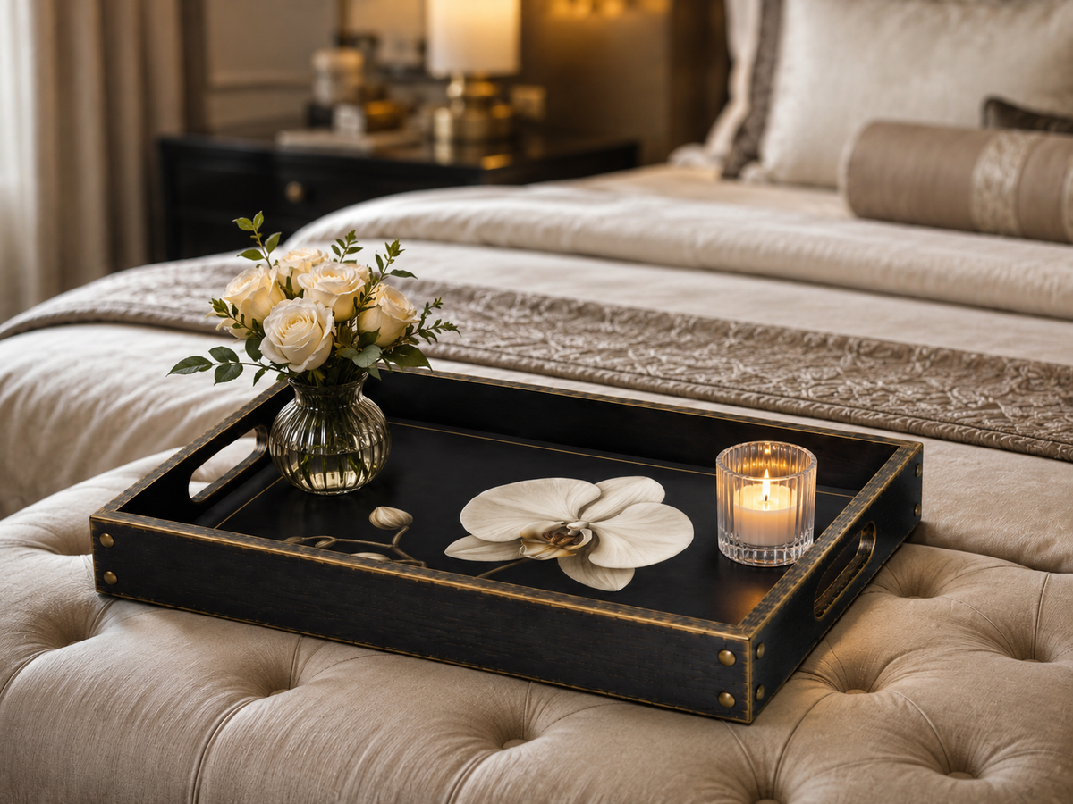 A rectangular black serving tray with gold trim and handles sits on a tufted beige ottoman in a softly lit bedroom. The tray holds a small glass vase with cream roses, a white orchid flower, a spoon, and a lit candle in a glass holder.