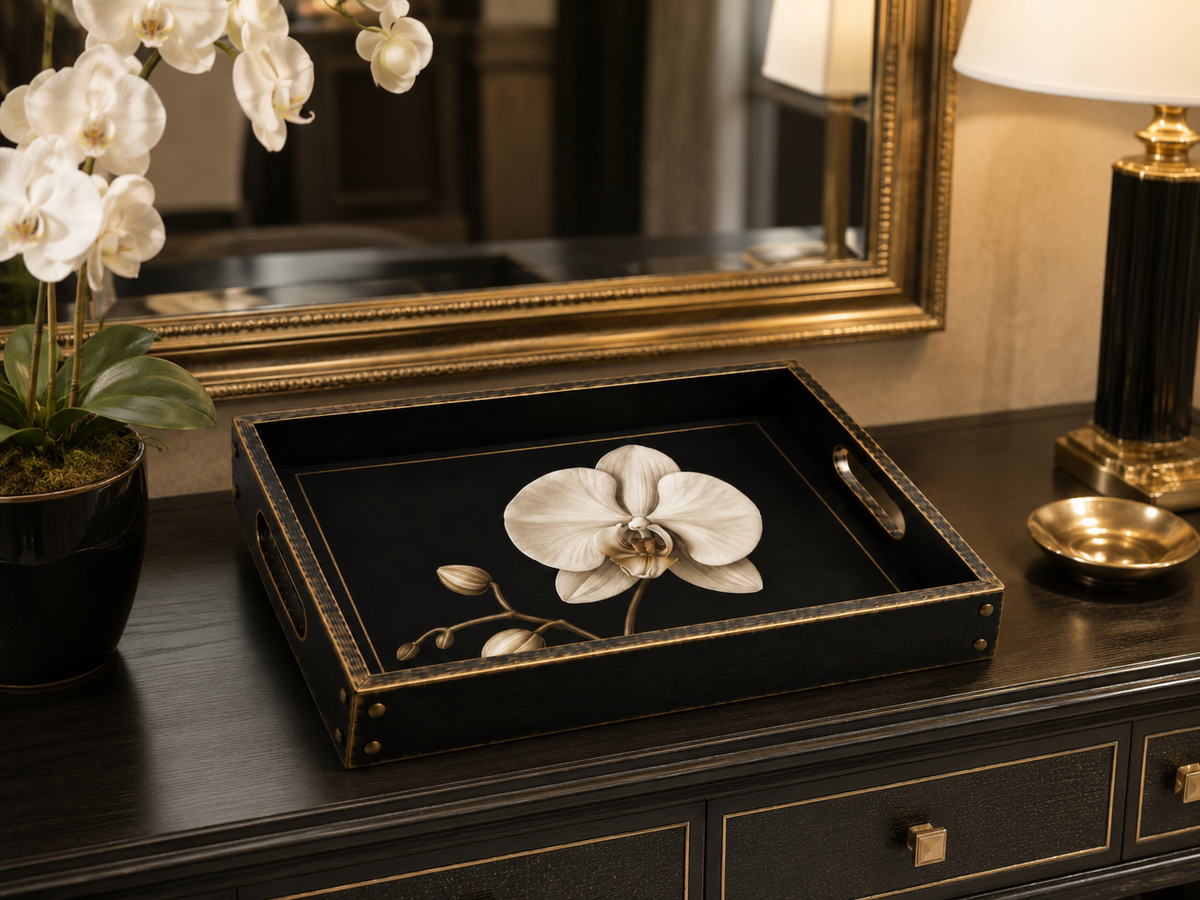 A black rectangular decorative tray with gold trim and a large white orchid illustration in the center, styled on a dark wood dresser next to a potted orchid and a lamp.