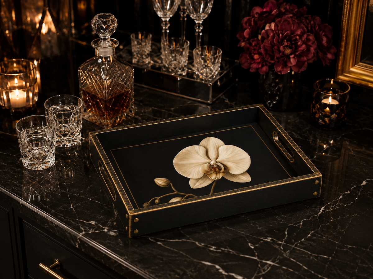 A black rectangular serving tray with gold trim and corner hardware, featuring a printed white orchid flower, styled on a dark marble countertop with glassware, a decanter, and candles.