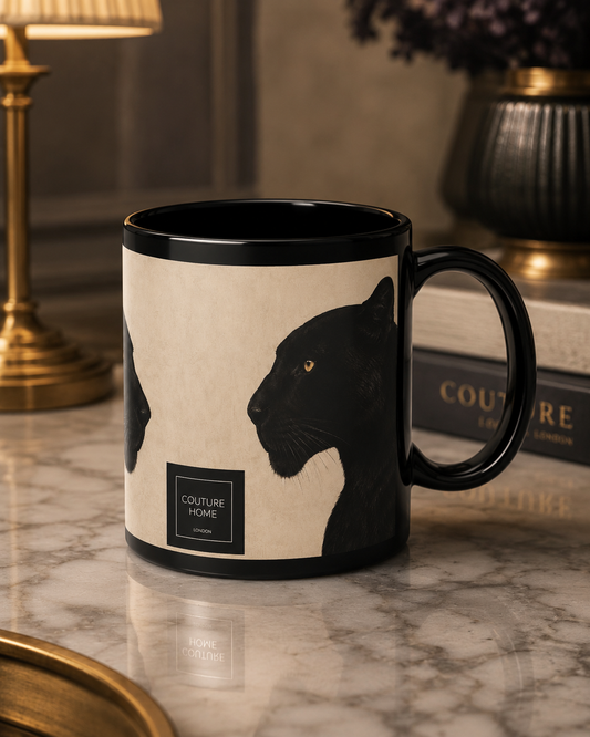 A black ceramic mug with a cream-colored wraparound print featuring black panther illustrations and a small 'COUTURE HOME LONDON' label, set on a marble surface with a lamp and books in the background.