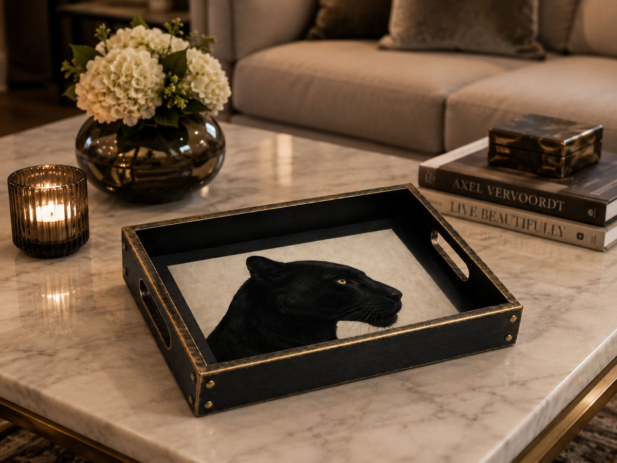 A black rectangular decorative tray with gold trim and cutout handles sits on a white marble coffee table in a styled living room. The tray features a black panther illustration on a light background.