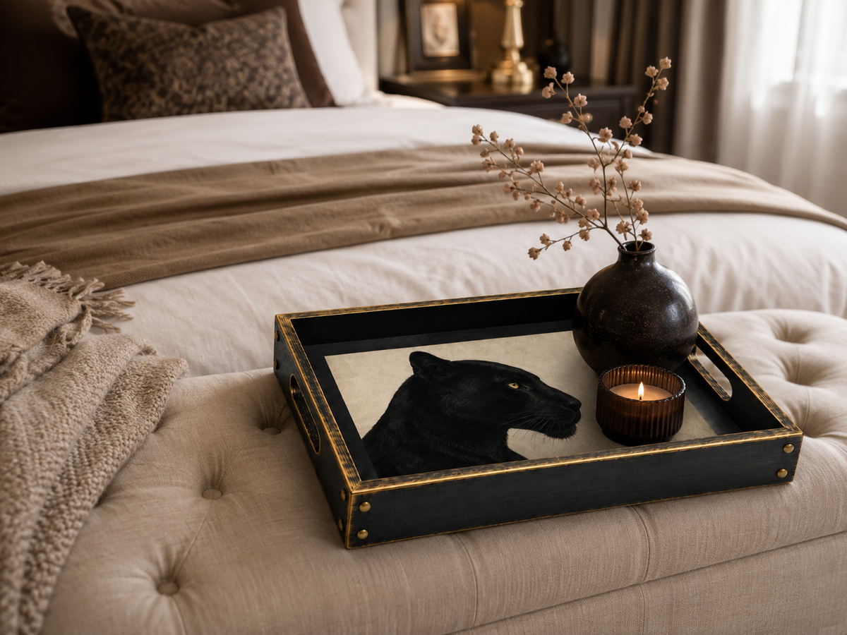 A black rectangular decorative tray with gold trim and a panther illustration, styled on a tufted beige bench in a bedroom setting with a black vase and candle.