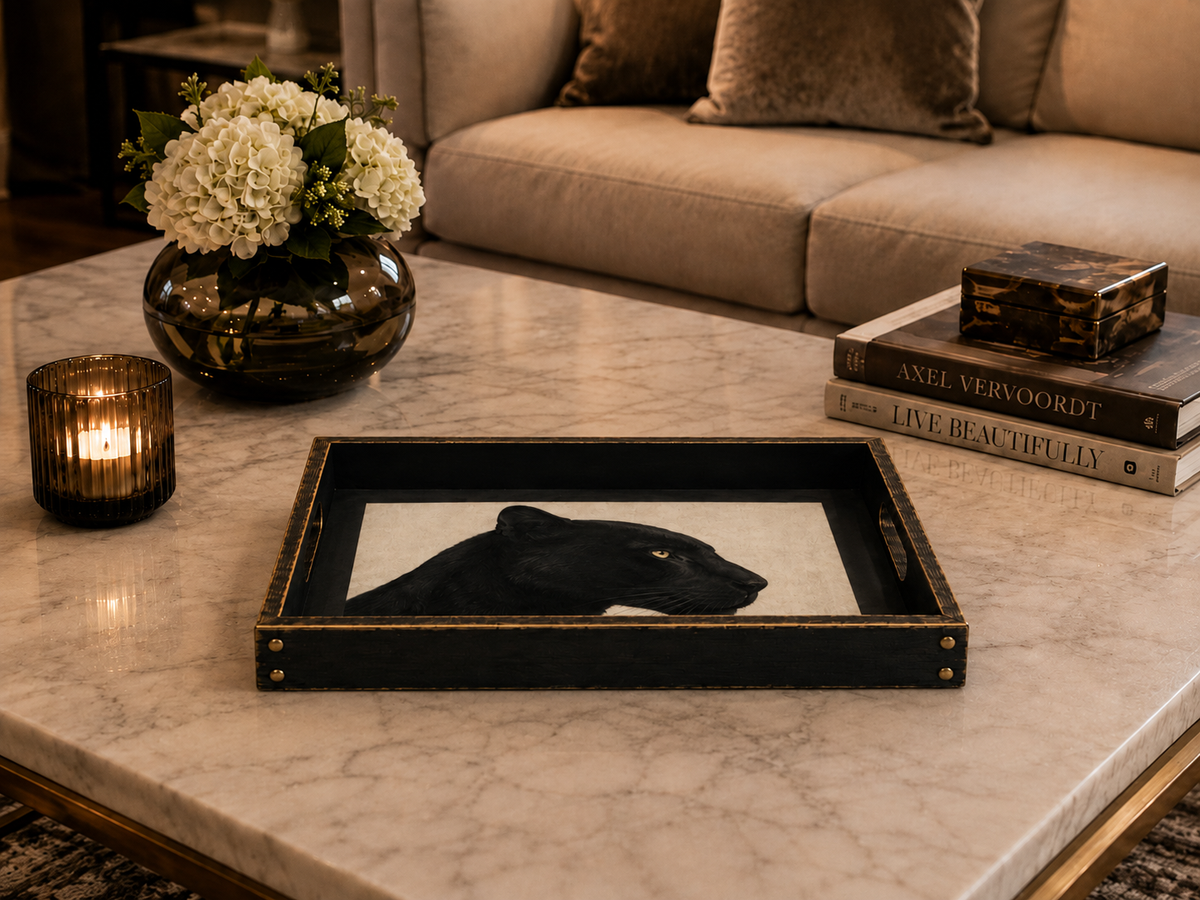 A rectangular black tray with gold trim and a black panther illustration on a marble coffee table in a living room setting, styled with a candle, flowers, and books.